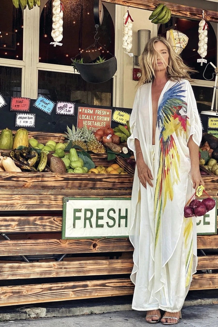 Kimono blanc avec imprimé perroquet coloré porté par une femme devant un étal de marché rempli de fruits et légumes frais. Nos kimonos pour un style unique et estival.