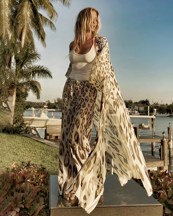 Femme blonde portant un kimono à motifs léopard et un pantalon assorti, posant devant un paysage tropical avec des palmiers et un quai. Vente de kimonos en ligne.