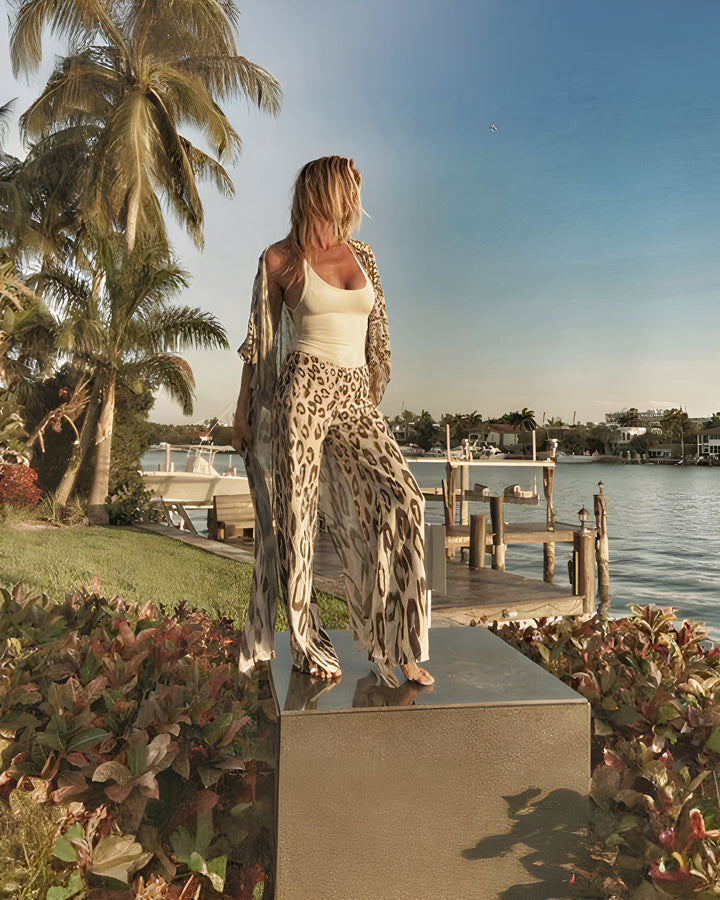Femme portant un kimono imprimé léopard et un pantalon assorti, regardant l'océan au coucher du soleil. Vêtements de plage et de style, vente de kimonos en ligne.