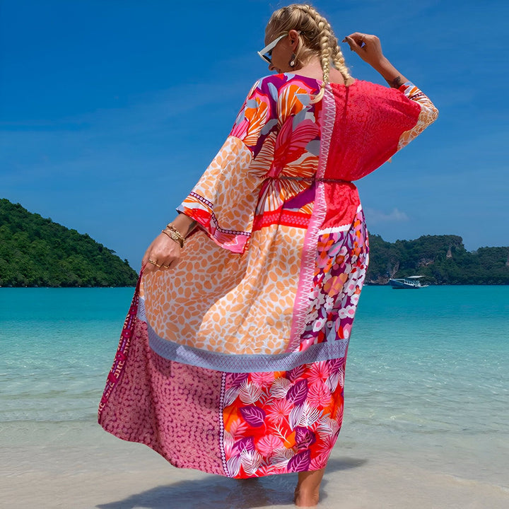 Femme blonde tressée, dos tourné, robe longue colorée à motifs floraux et géométriques, plage de sable blanc, eau turquoise, îles verdoyantes en arrière-plan. Style bohème chic, vacances ensoleillées, mode estivale.