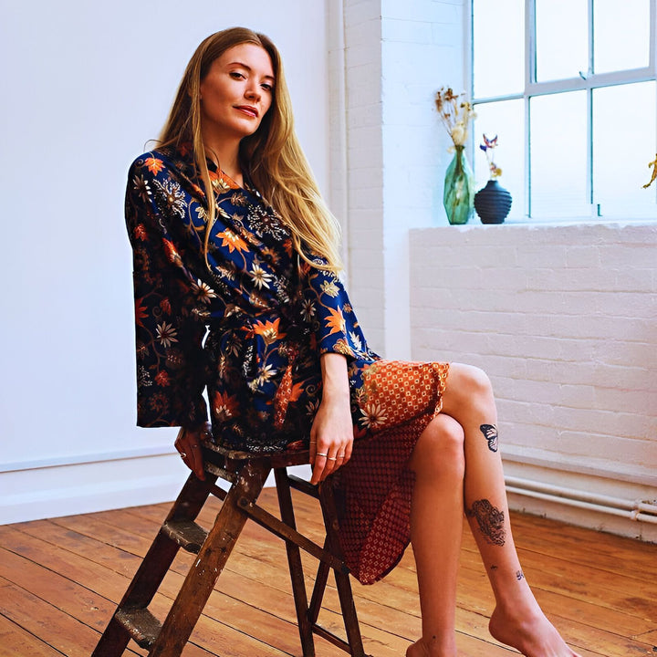 Une femme blonde, assise sur un escabeau près d'une fenêtre, porte une robe à motifs floraux bleus et oranges. Elle a des tatouages sur la jambe et regarde la caméra. Style décontracté et lumineux.
