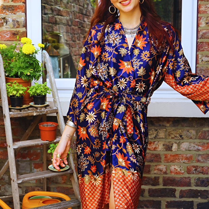Femme souriante portant un kimono bleu et orange à motifs floraux. Échelle en bois avec des plantes en pot en arrière-plan, devant un mur de briques. Bijoux argentés, créoles. Style bohème.