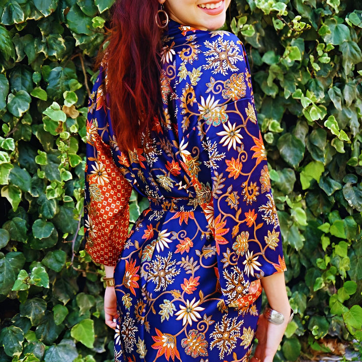 Femme souriante portant un kimono bleu à motifs floraux orange et blancs devant un mur de lierre. Détail des manches rouges à motifs. Cheveux roux lâchés et boucles d'oreilles dorées.