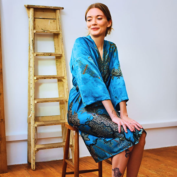 Femme souriante assise, portant un kimono bleu à motifs. Échelle en bois à l'arrière-plan. Découvrez notre collection de kimonos élégants.