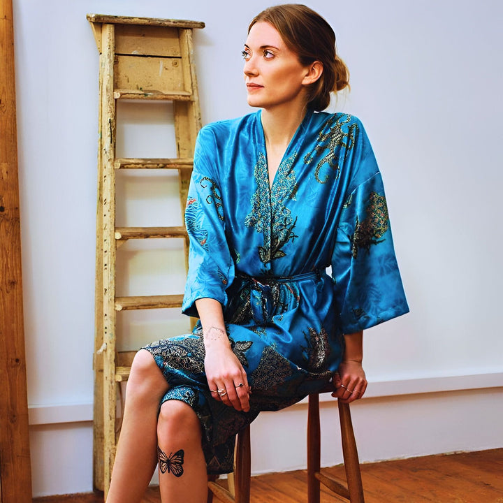 Femme assise portant un kimono bleu orné de motifs floraux. Fond blanc avec une échelle en bois rustique. Vêtements de détente élégants. Kimonos en vente.