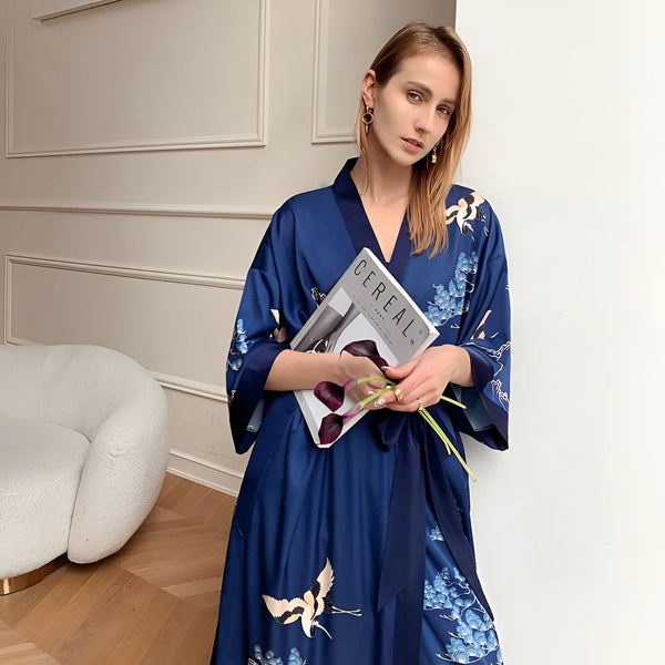 Femme portant un kimono bleu orné de motifs floraux et de grues, tenant un magazine et des fleurs. Vêtements de nuit élégants. Boutique de kimonos en ligne.