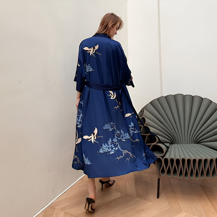 Femme portant un kimono bleu marine élégant avec des motifs de grues et de fleurs. Kimono long et fluide avec une ceinture assortie. Vêtement traditionnel japonais revisité. Idéal pour se détendre avec style. Découvrez notre collection de kimonos uniques.