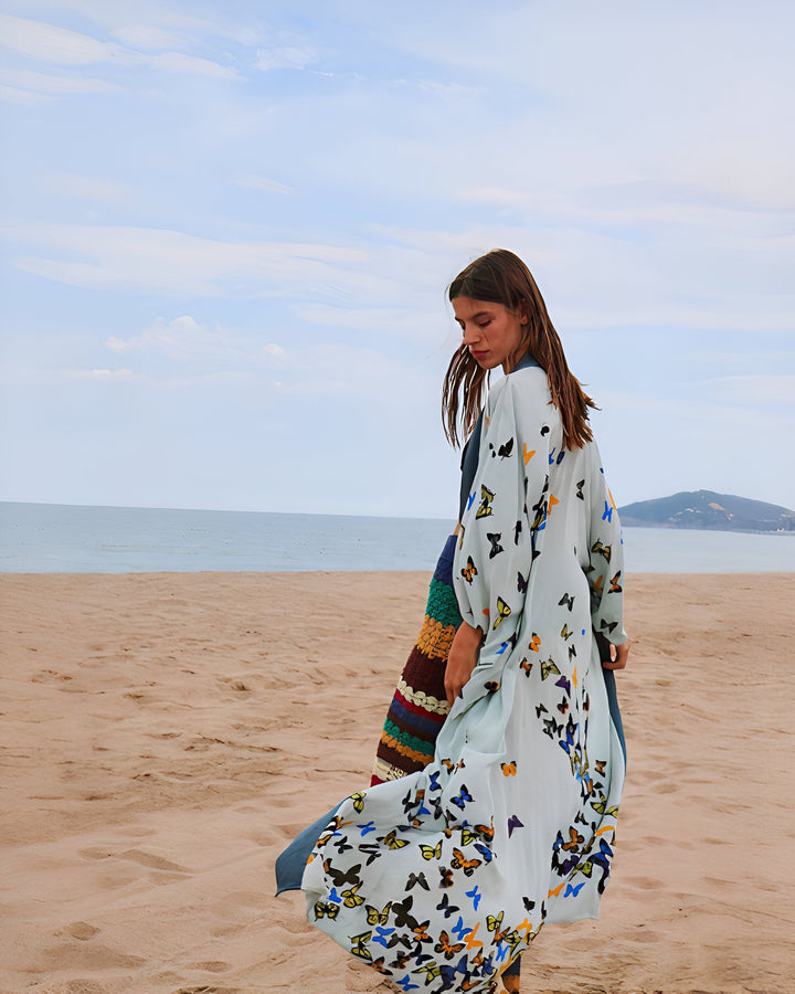 Femme portant un kimono bleu clair à motif papillons sur la plage. Kimono long et fluide, idéal pour l'été. Découvrez notre collection de kimonos tendance.