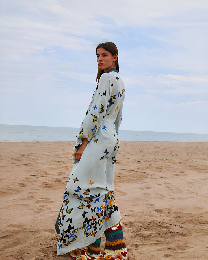 Femme portant un kimono long imprimé papillons sur une plage de sable. Kimonos élégants et confortables disponibles à l'achat.