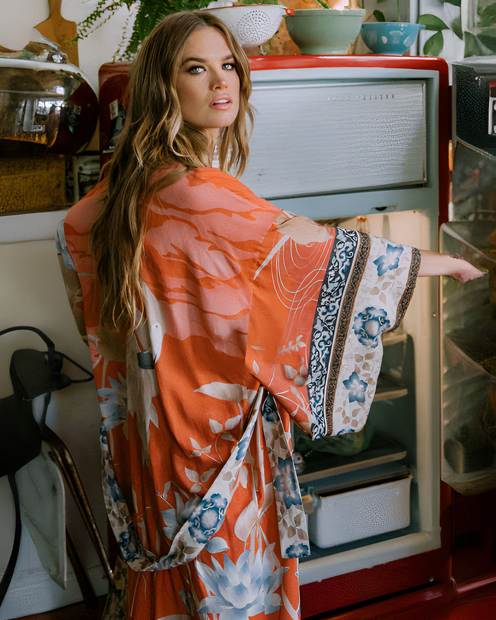 Femme portant un kimono orange à motifs floraux devant un réfrigérateur vintage. Élégance et confort pour la maison. Découvrez notre collection de kimonos uniques.
