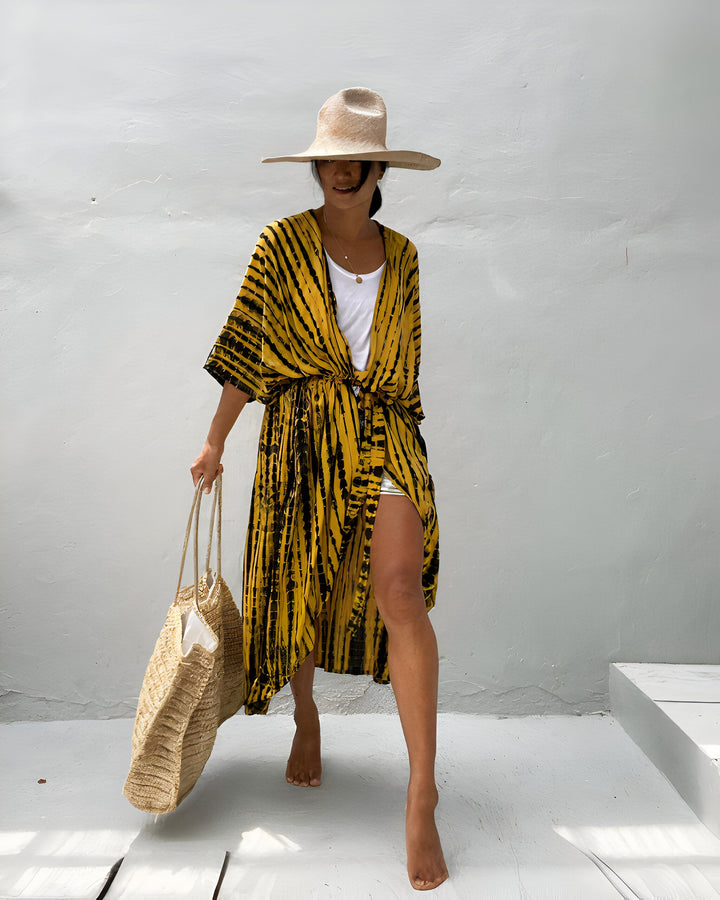 Femme portant un kimono jaune à rayures noires, un chapeau de paille et un sac en paille, pieds nus. Style estival décontracté, mode vacances.