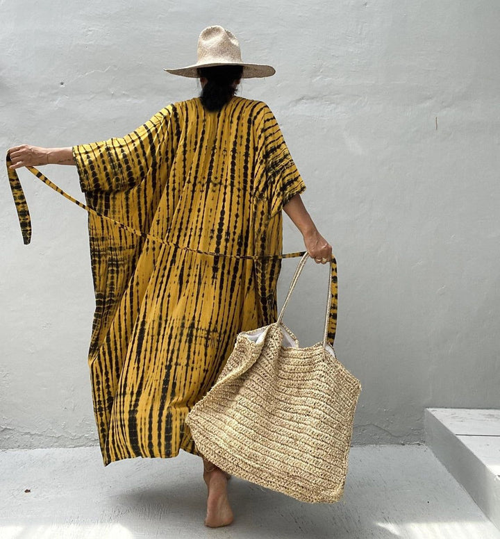 Femme vue de dos portant une robe jaune à rayures noires, un chapeau de paille et un grand sac en paille. Style bohème, été.