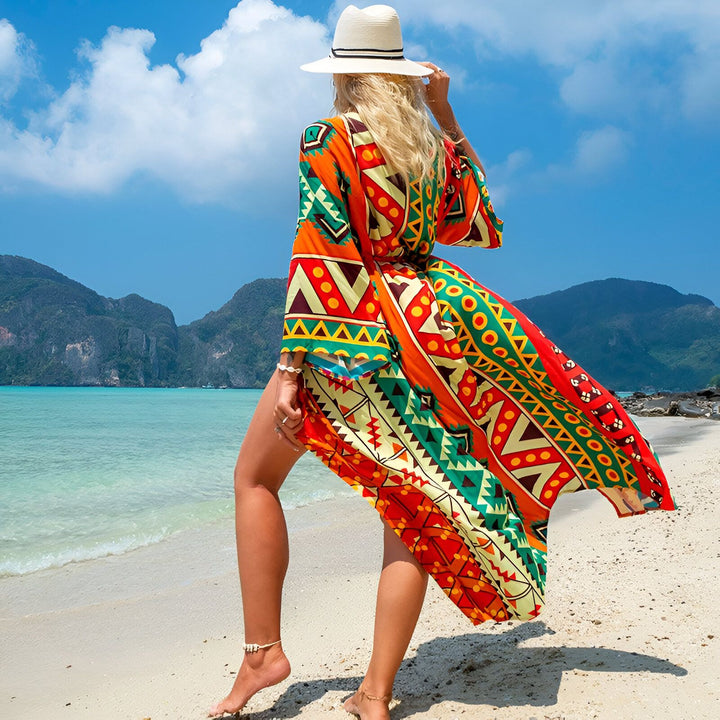 Femme blonde, dos tourné, portant un kimono coloré à motifs sur une plage tropicale. Kimono long et ample, chapeau de paille. Notre collection de kimonos pour un style unique.