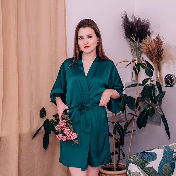 Femme en kimono de soie vert émeraude, tenant des roses roses. Vêtements d'intérieur élégants pour femme. Découvrez notre collection de kimonos de luxe.