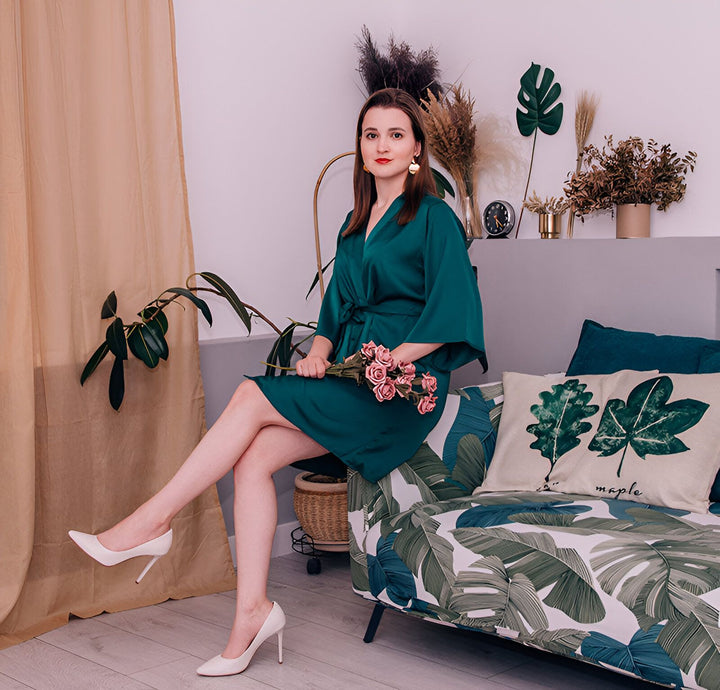 Femme élégante en kimono vert émeraude, assise sur un canapé à motifs végétaux. Elle tient des roses roses. Vente de kimonos en ligne.