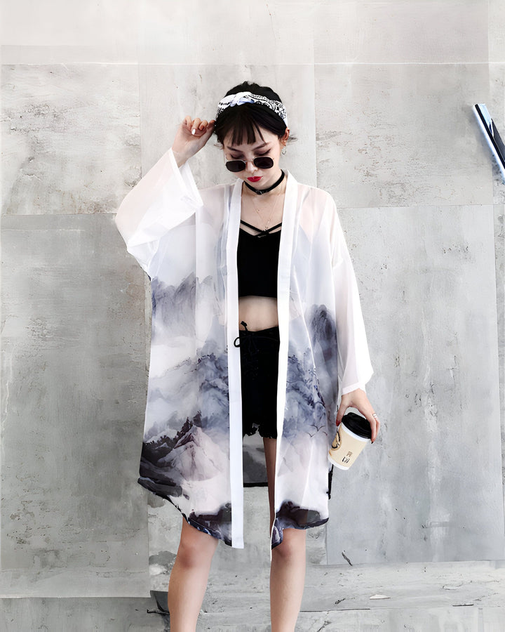 Femme portant un kimono blanc transparent avec motif de montagnes, lunettes de soleil rondes et bandeau. Kimono tendance pour un look bohème chic. Découvrez notre collection de kimonos uniques.