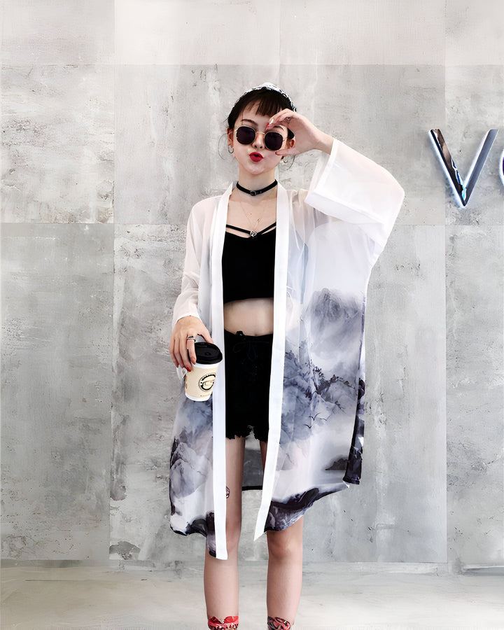 Femme portant un kimono blanc imprimé avec des motifs de montagne. Elle porte un haut noir court et des shorts noirs. Look décontracté avec lunettes de soleil et café à la main. Kimonos tendance pour toutes les occasions.