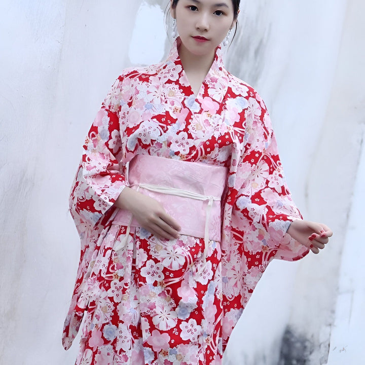 Femme portant un kimono traditionnel japonais rouge à motifs floraux roses et blancs, accessoirisé d'une ceinture obi rose pâle. Vêtements japonais authentiques pour un style unique.