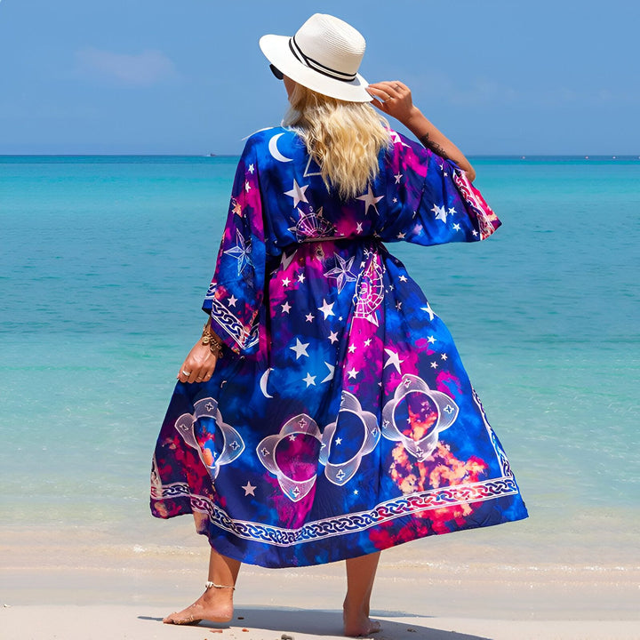 Femme portant un kimono long bleu avec des motifs cosmiques (lunes, étoiles) sur une plage de sable fin. Vêtement de plage élégant et coloré. Notre collection de kimonos.