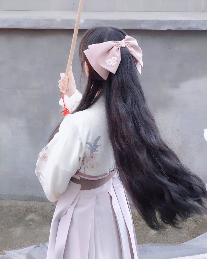 Femme portant un kimono rose et blanc avec motif floral délicat, vue de dos. Elle a un grand nœud rose dans les cheveux et tient un parapluie. Vêtements traditionnels japonais en vente.