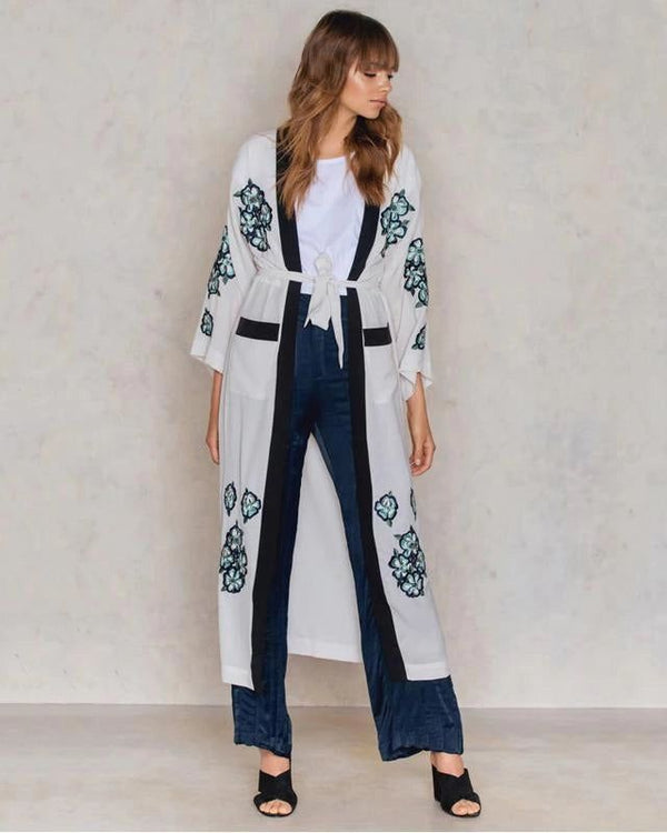Femme portant un kimono blanc brodé de motifs floraux, avec une bordure noire. Elle porte un pantalon bleu foncé et des sandales noires à talons.