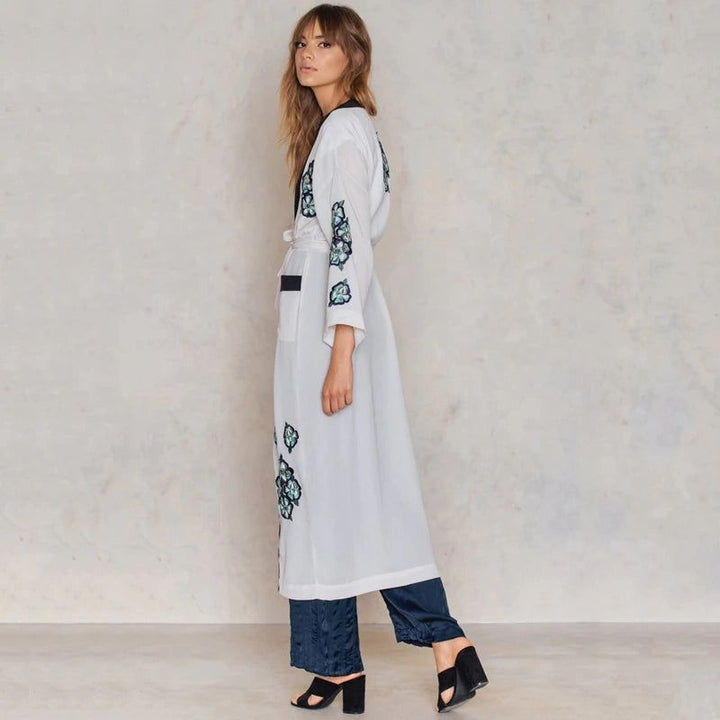 Femme portant un kimono blanc long avec des broderies florales bleues, un pantalon bleu ample et des talons noirs. Style bohème chic.