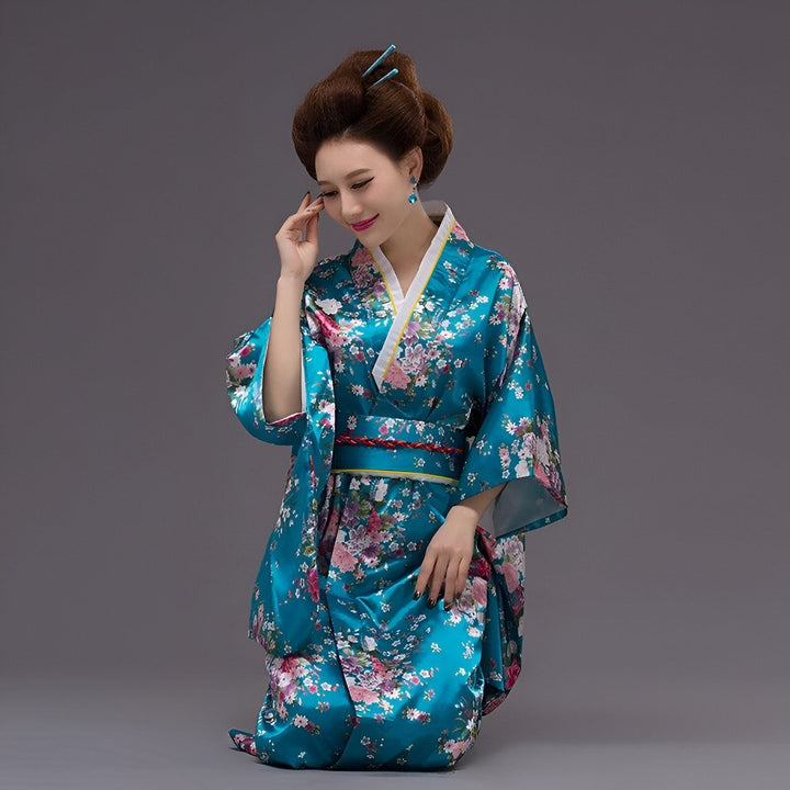 Jeune femme asiatique à genoux, vêtue d'un kimono bleu à motifs floraux roses et blancs. Elle porte des épingles à cheveux bleues et des boucles d'oreilles assorties, coiffée de façon traditionnelle. Elle sourit légèrement.