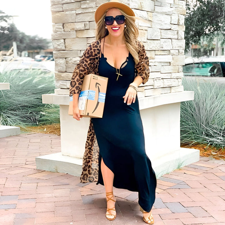 Femme souriante portant un kimono léopard, une robe noire, un chapeau et des lunettes de soleil, tenant un colis Amazon. Kimonos tendance pour toutes les occasions.