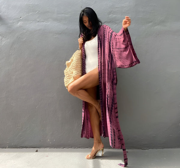 Femme portant un kimono long violet tie-dye, maillot de bain blanc et talons hauts dorés. Sac de plage en paille. Kimonos tendance à acheter en ligne.