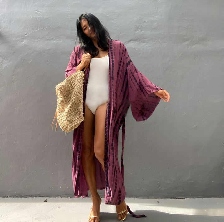 Femme portant un kimono long tie-dye violet, maillot de bain blanc, sac en paille et talons dorés. Vêtements de plage, style décontracté, boutique de kimonos en ligne.