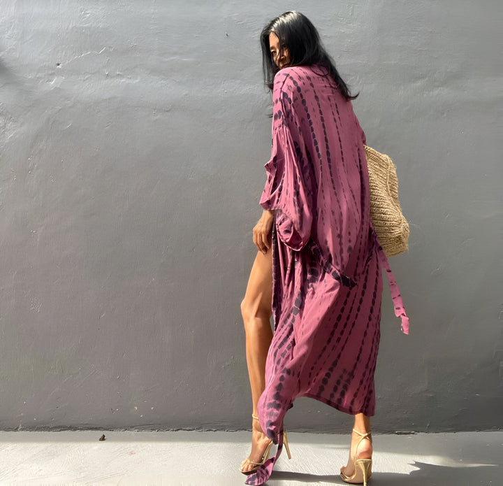 Femme portant un kimono tie-dye rose et noir avec un sac en paille, talons dorés. Vêtements style kimono pour femme.