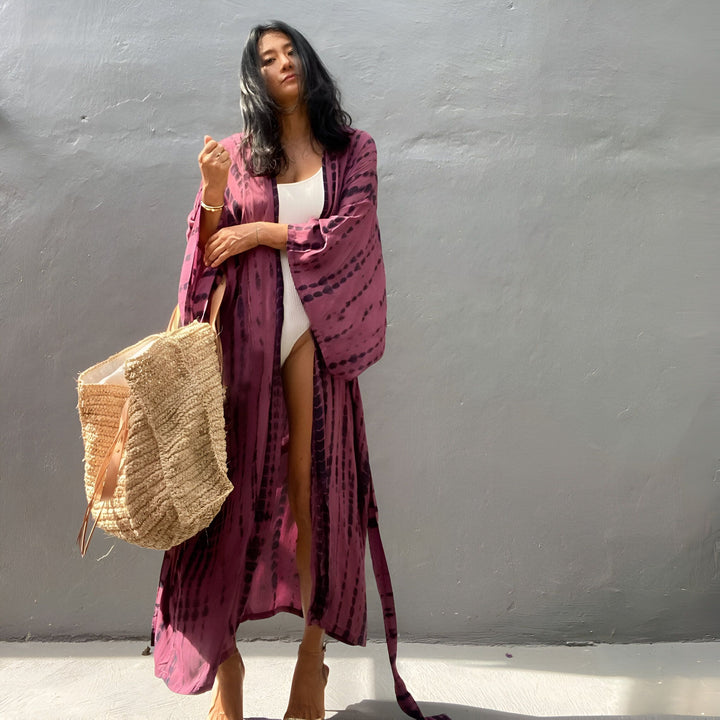Femme portant un kimono long tie-dye violet et noir avec un maillot de bain blanc, tenant un sac de plage en paille. Kimonos de notre collection.
