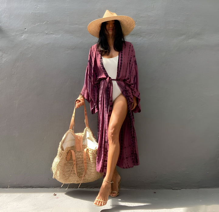 Femme élégante portant un kimono tie-dye violet, un maillot de bain blanc et un chapeau de paille. Sac de plage en paille. Découvrez notre collection de kimonos tendance.