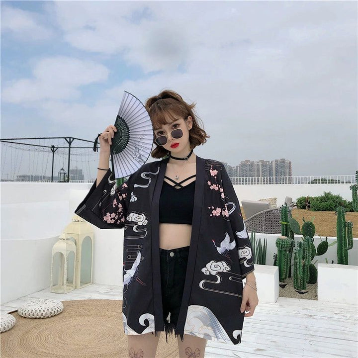 Femme portant un kimono noir à motifs de grues et de fleurs de cerisier, tenant un éventail. Vêtements de style japonais à la mode. Kimonos disponibles à l'achat.