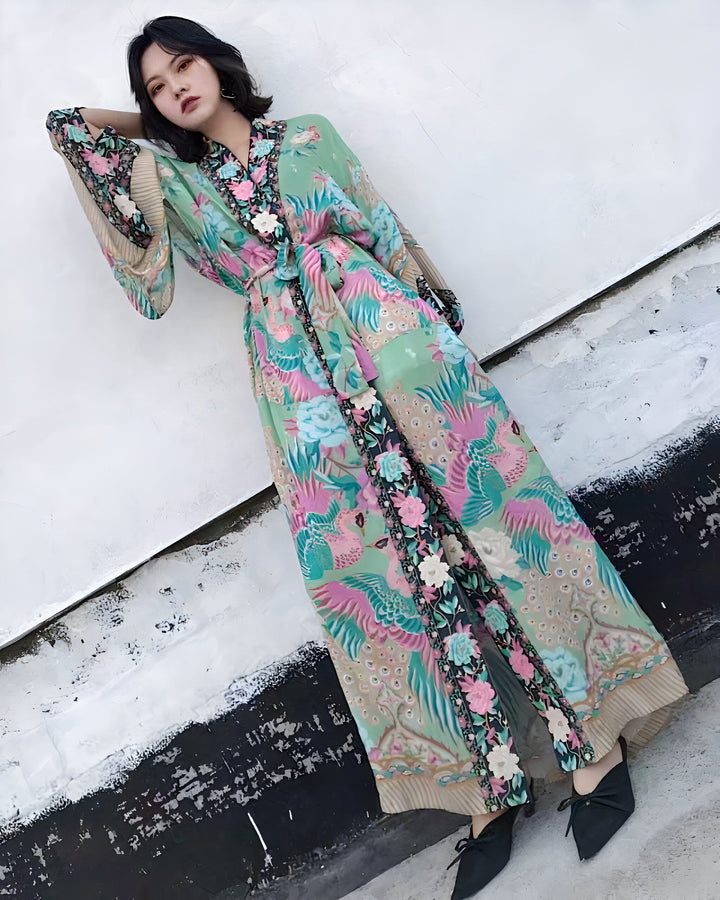 Femme portant un long kimono vert avec des motifs floraux et d'oiseaux roses et bleus. Vêtement de style japonais, parfait pour la détente à la maison ou comme tenue élégante. Découvrez notre collection de kimonos uniques.