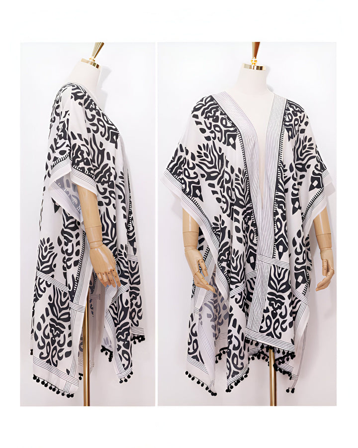 Kimono blanc à motifs noirs présenté sur mannequin. Vêtement d'extérieur ample avec manches courtes et détails pompons. Idéal pour un look décontracté et élégant.
