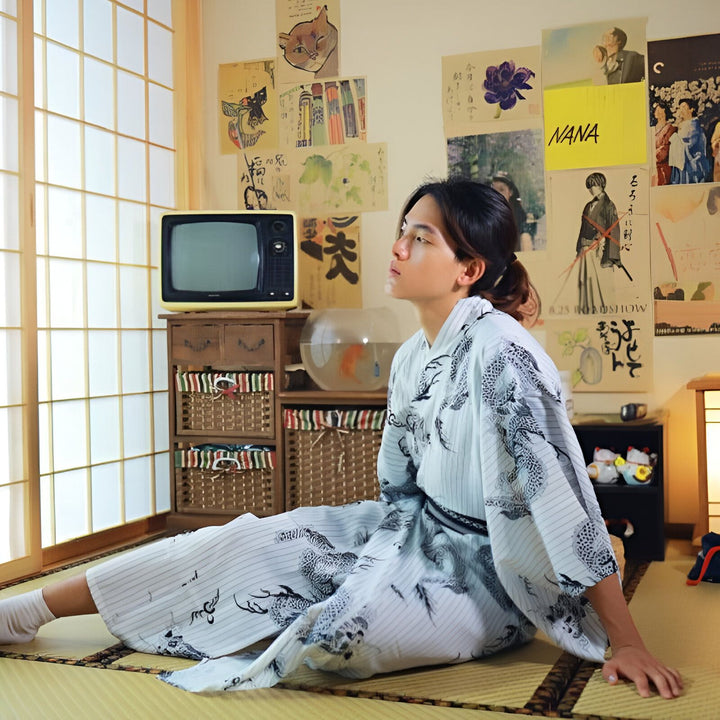 Homme assis sur un tatami, portant un kimono blanc à motifs de dragons noirs. Décor japonais traditionnel avec affiches, TV vintage et aquarium. Découvrez notre collection de kimonos authentiques.