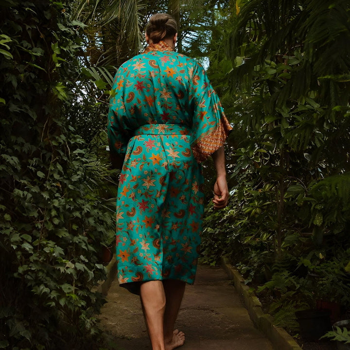 Homme vu de dos, portant un kimono turquoise à motifs floraux orange et rouges, se promenant dans une serre luxuriante. Vente de kimonos en ligne.