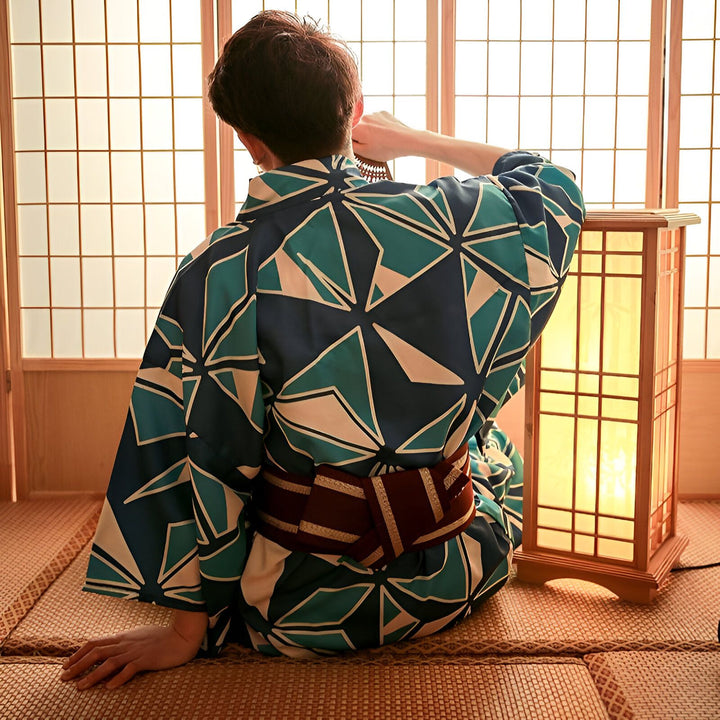 Homme portant un kimono japonais bleu et vert à motifs géométriques. Ceinture obi bordeaux. Ambiance zen et traditionnelle avec tatami et lanterne japonaise. Kimonos pour hommes disponibles à l'achat.