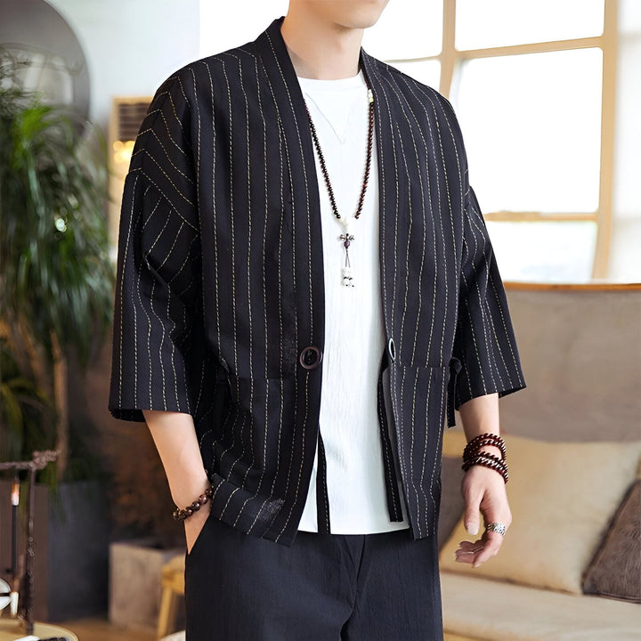 Homme portant un kimono noir à rayures blanches avec un t-shirt blanc et un collier, et un bracelet. Vêtements décontractés pour hommes avec style moderne.