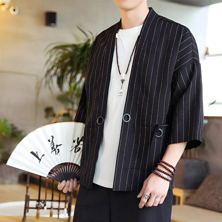 Homme en veste kimono noire à rayures, tenant un éventail blanc avec écriture asiatique. Collier à perles, bracelets rouges et bague argentée. Style décontracté et élégant.