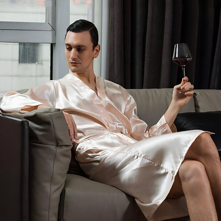 Homme portant un kimono en soie couleur chair, assis sur un canapé et tenant un verre de vin rouge. Kimonos élégants pour hommes, disponibles à l'achat. Confort et luxe dans un vêtement traditionnel revisité.