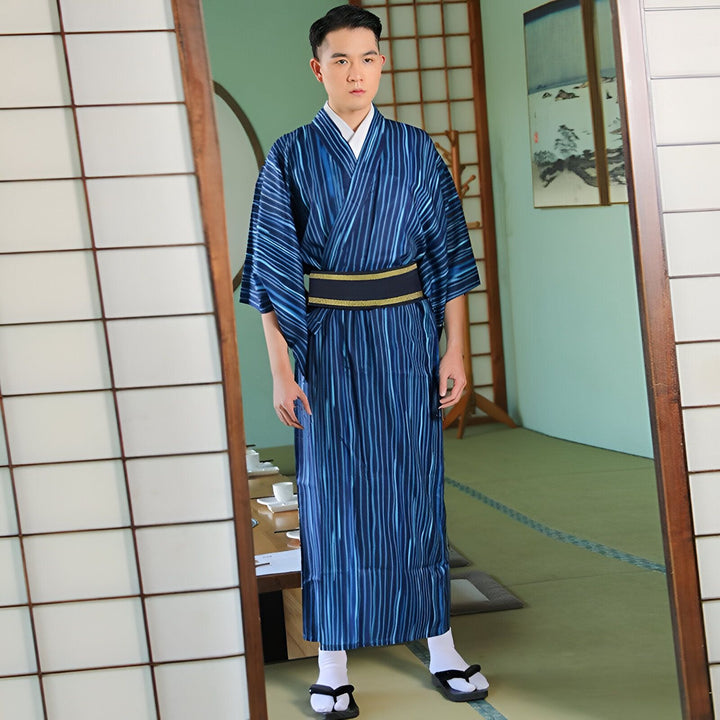 Homme portant un kimono traditionnel japonais bleu à rayures, avec une ceinture obi noire et or, dans un intérieur japonais authentique. Découvrez notre collection de kimonos uniques.