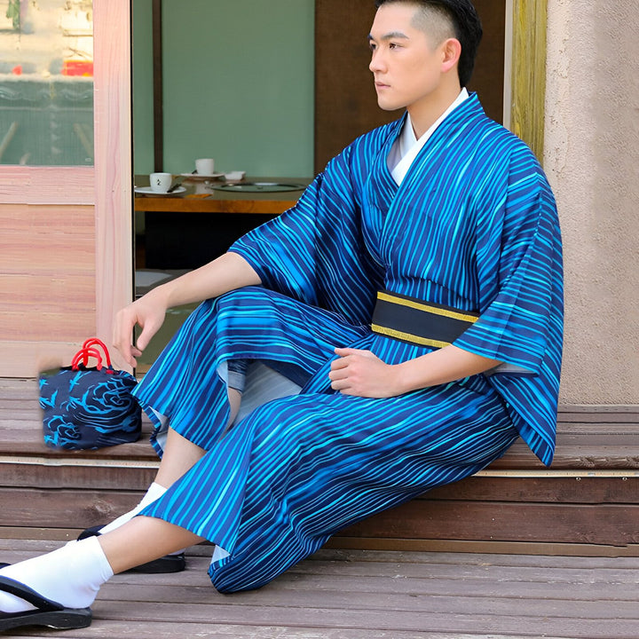 Homme en kimono rayé bleu, assis sur des marches en bois. Kimono traditionnel japonais, ceinture noire, chaussettes blanches et sandales. Sac bleu avec motifs. Vêtements japonais, style estival.