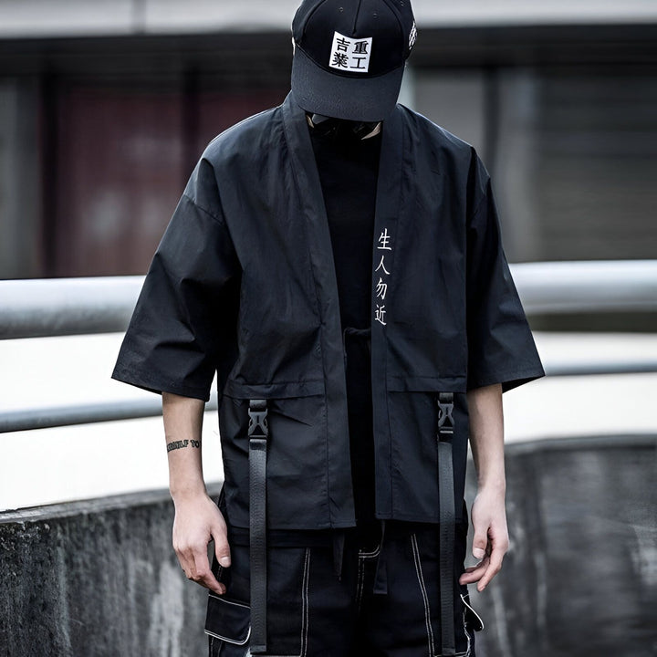 Homme portant un kimono noir moderne avec des caractères japonais brodés. Veste ample avec poches et sangles. Style urbain et décontracté. Idéal pour un look tendance et original. Découvrez notre collection de kimonos uniques.