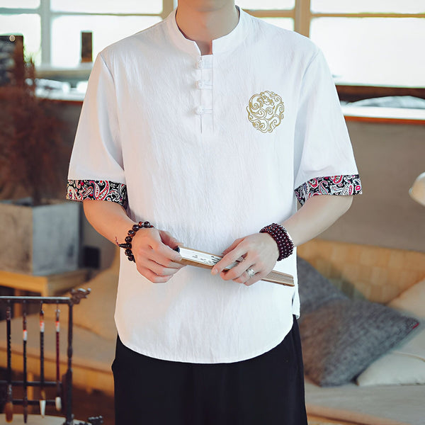 Homme portant une chemise blanche à manches courtes avec motifs floraux brodés, col mao et poignets décorés. Il tient un éventail en bois et porte des bracelets en perles.