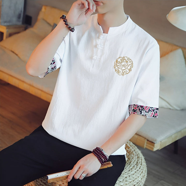 Homme portant un chemisier blanc à manches courtes avec motif brodé et poignets décorés, style asiatique, assis. Il a un bracelet en perles au poignet et tient un éventail.