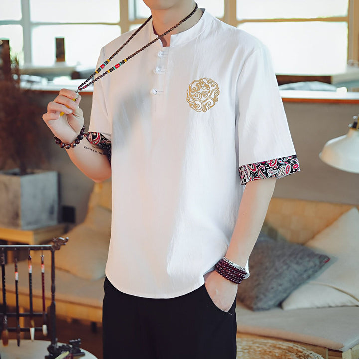 Homme portant une chemise blanche à manches courtes avec motif brodé doré et bordures imprimées, pantalon noir. Collier de perles et bracelets. Style asiatique, décontracté.