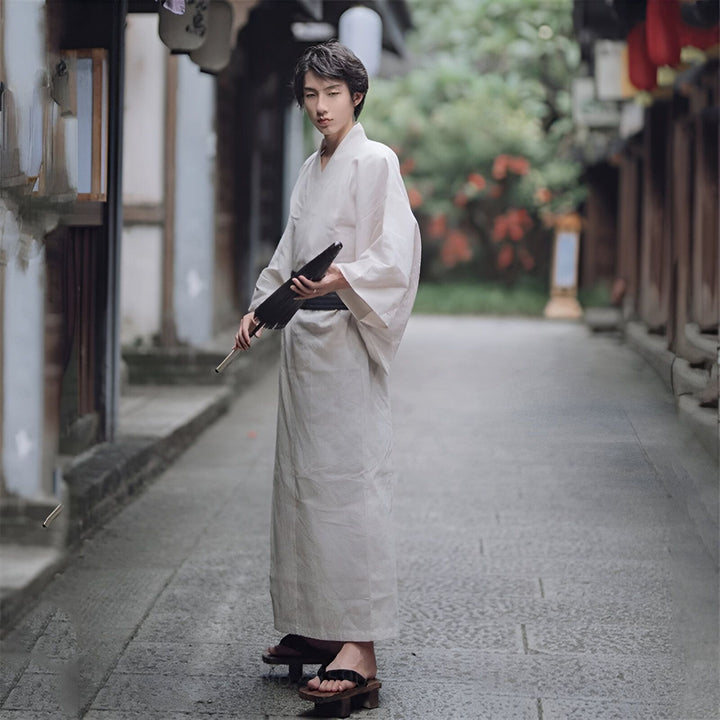 Un homme en kimono blanc, tenant un parapluie japonais, dans une rue pittoresque. Découvrez notre collection de kimonos traditionnels et modernes.