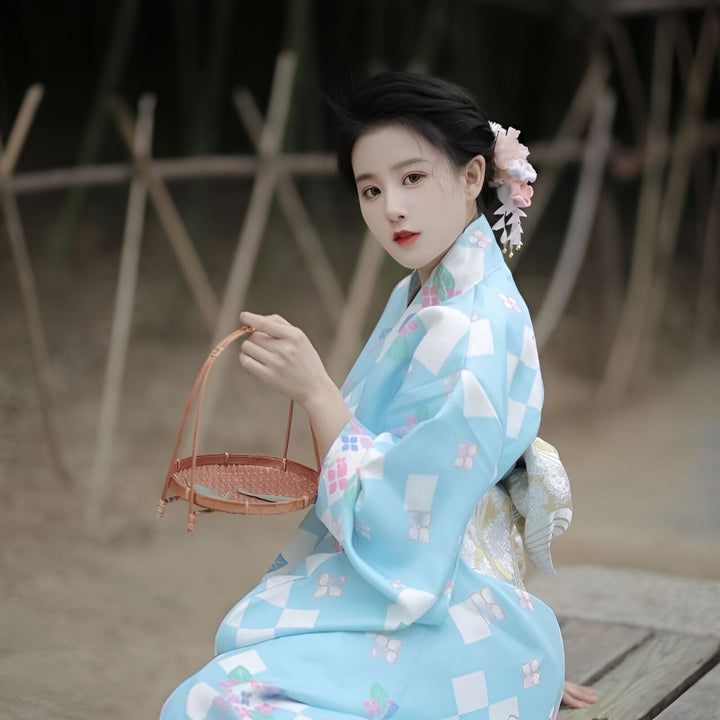 Femme asiatique élégante en kimono bleu à motifs géométriques et floraux. Elle porte une fleur dans les cheveux et tient un panier en bambou. Kimonos à la vente.